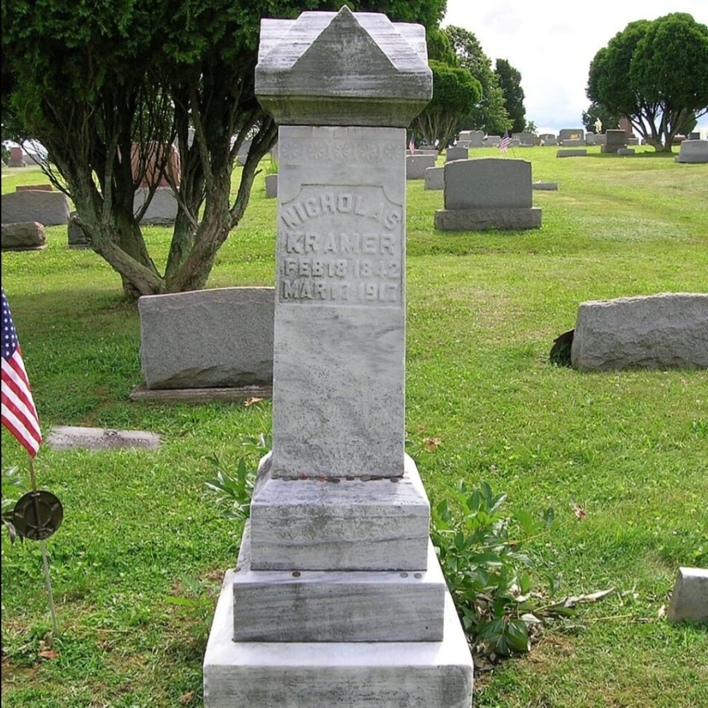 Tall marble gravestone of Nicholas Kramer at Evans City Cemetery, surrounded by grass, trees, and nearby markers.