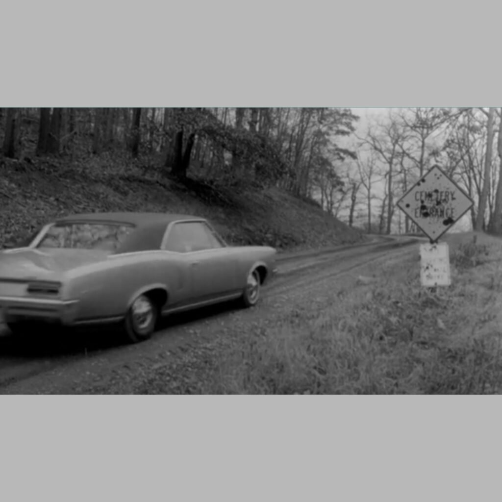 Black‑and‑white screenshot of the original “Cemetery Entrance” sign from Night of the Living Dead showing the rural road leading into Evans City Cemetery.